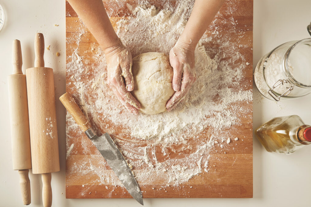 top view on white table with isolated wooden board with knife two rolling pins bottle olive oil transparent jar with flour woman hands hold prepared dough for pasta or dumplings